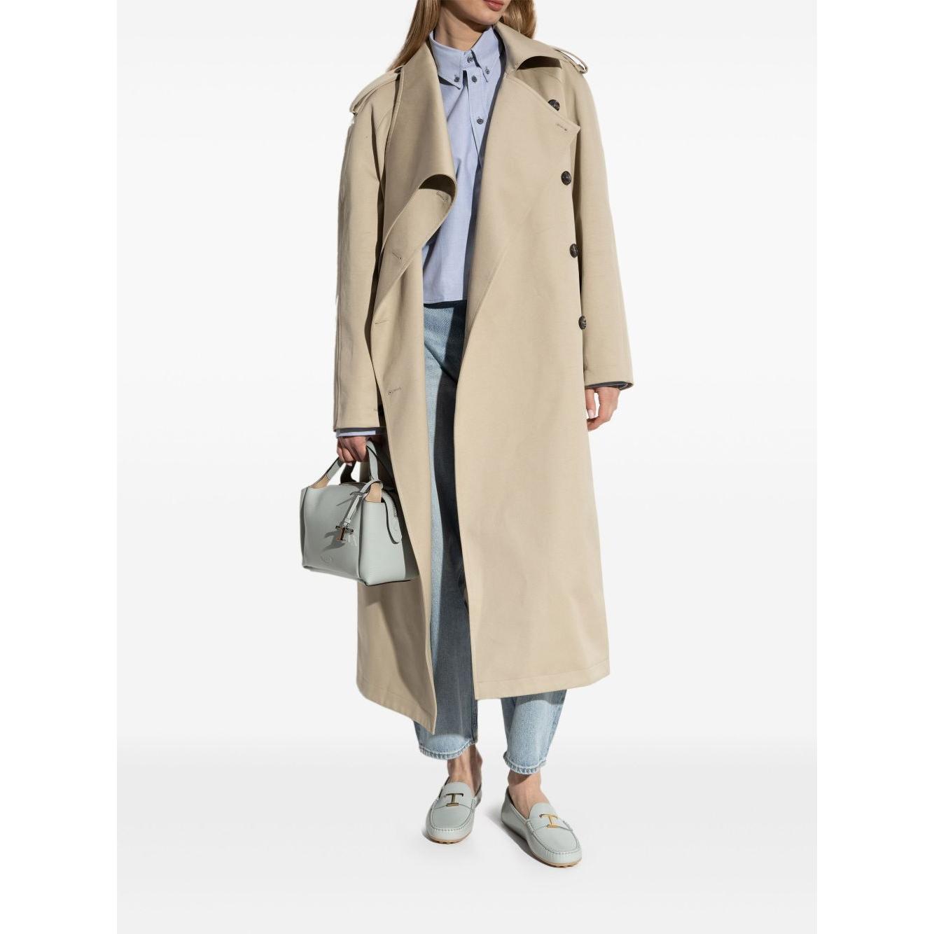 Person wearing a beige trench coat with light blue flat shoes and holding a designer handbag.