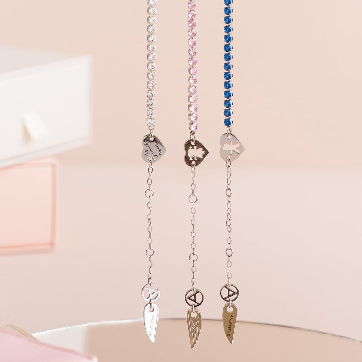 Elegant chain jewelry from AN JEWELS JEWELRY Mod. AL.BLIYMBL displayed on soft pink backdrop with delicate charms and colored gemstones.
