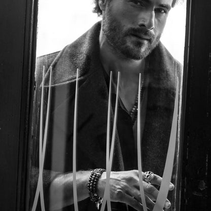 Man wearing chic attire and accessories, standing behind a glass door, showcasing stylish rings and bracelet with a thoughtful expression.