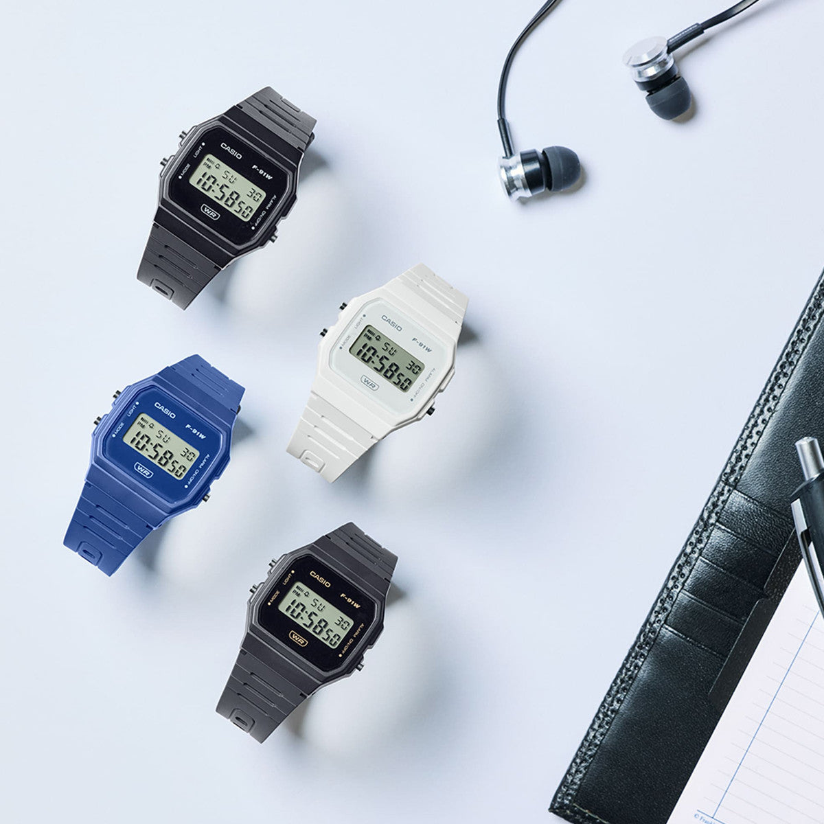 Four CASIO VINTAGE Mod. F-91 BIO RESIN STRAP - BLUE watches displayed with earphones and notepad for unisex style.