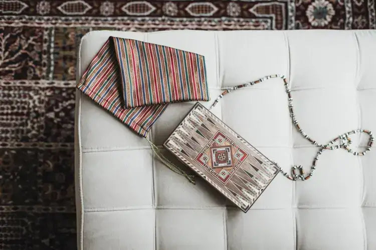 Decorative clutch bag with tribal pattern on a white cushion, accompanied by a striped fabric, on a patterned carpet background.