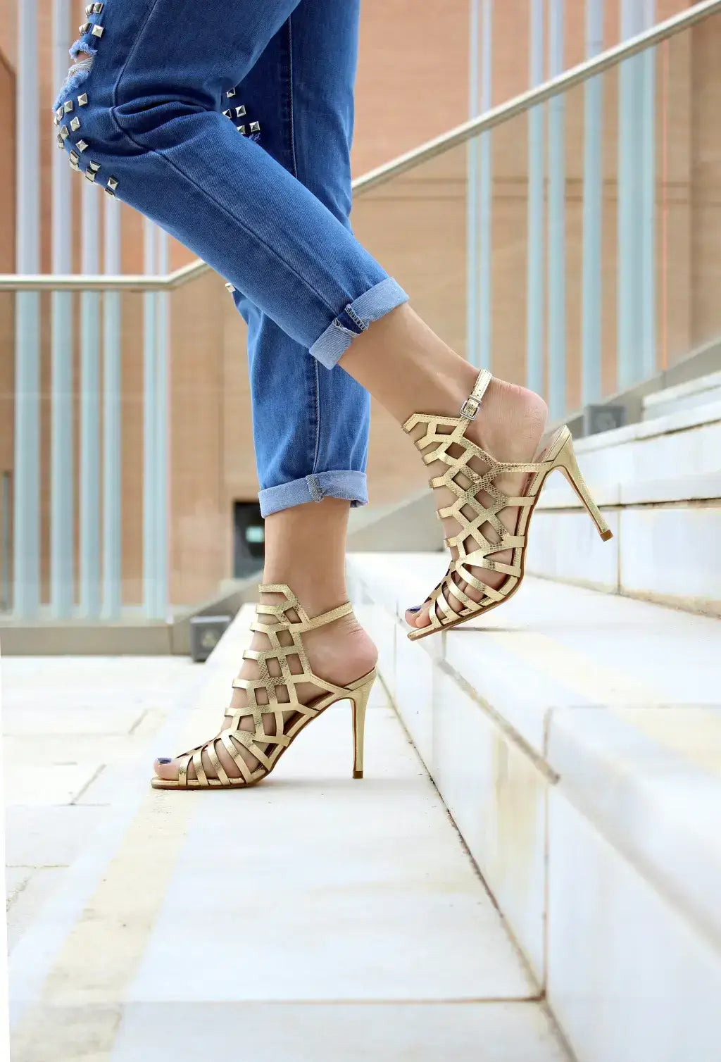 Woman wearing gold high heel sandals with jeans on stairs outside.