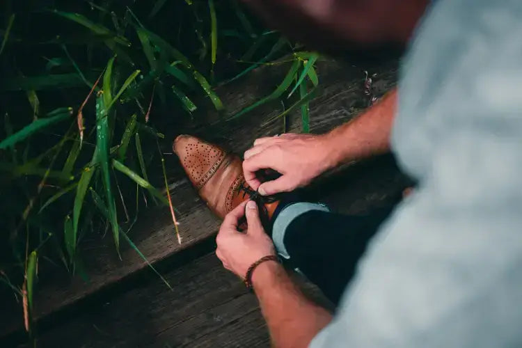 Man tying brown leather dress shoe near green leaves on wooden deck