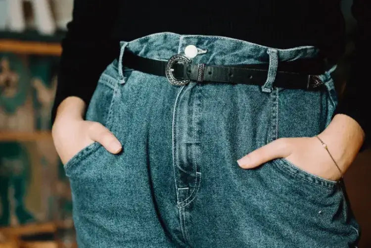 Person wearing blue denim jeans with hands in pockets and a black belt with a decorative buckle.